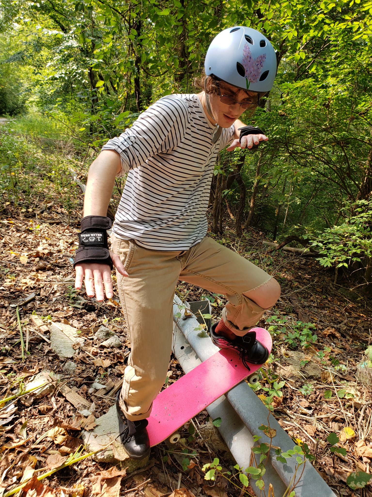 a horizontal black/white striped shirt, tan jeans that are ripped on the knee, a blue biking helmet with a lilac painted on it, wrist pads, a pink skateboard, and black hiking boots. im grinding on an abandoned road railing
